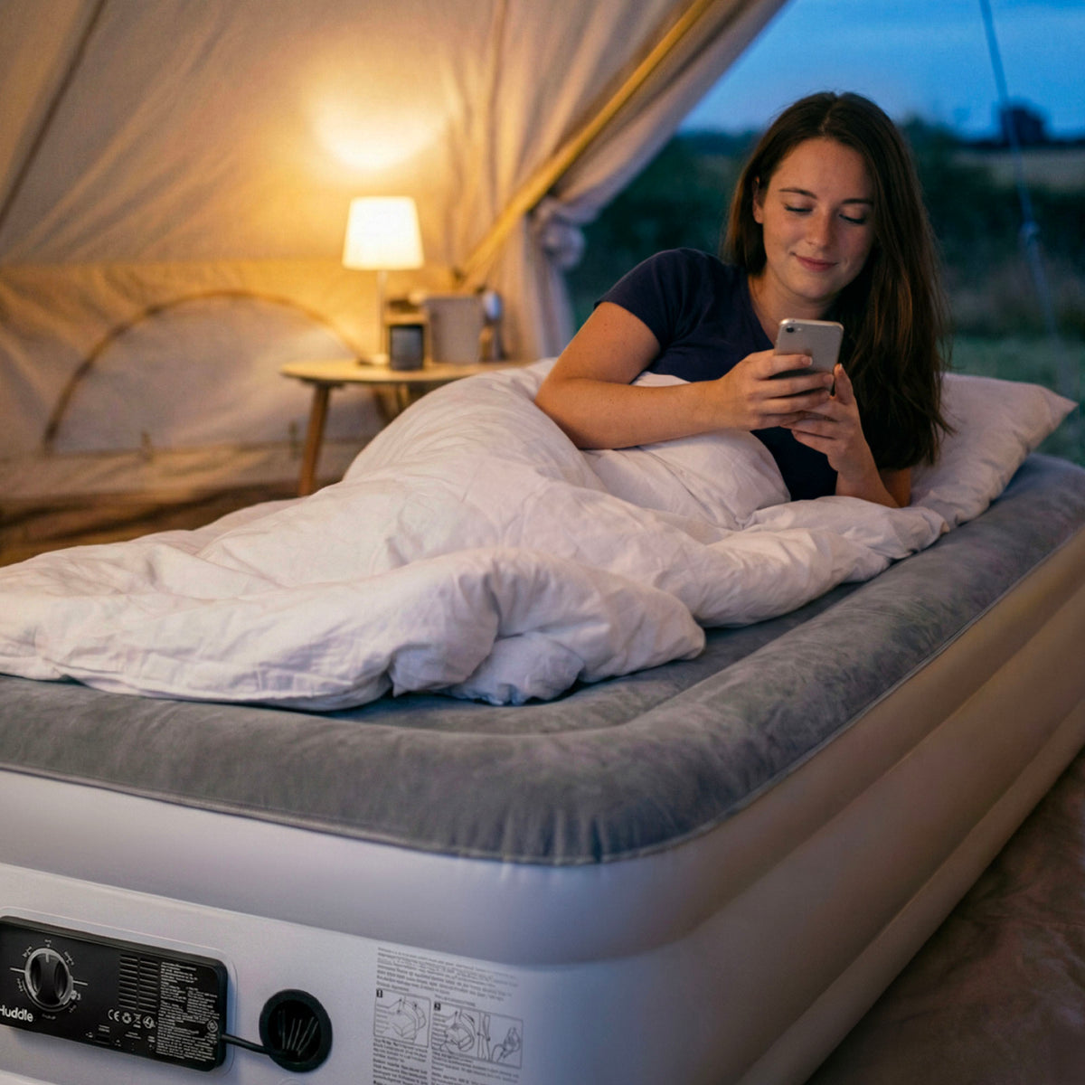 Woman relaxes on a Huddle air mattress inside a tent, using her phone; dual pump is visible at the foot