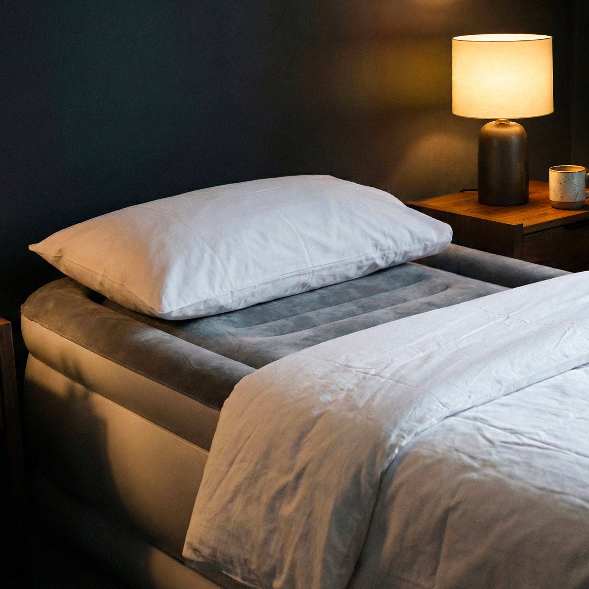 Single air mattress dressed with white bedding and a pillow in a warmly lit bedroom beside a lamp and nightstand